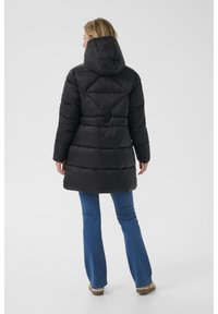 Abrigo negro acolchado con capucha, que presenta un diseño acolchado y mangas largas. La parte trasera tiene un acento triangular y se combina con unos vaqueros de campana azules.