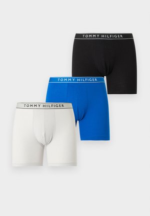 Homme torse nu portant un boxer noir Tommy Hilfiger, debout pieds nus devant un fond gris clair uni, regardant vers la gauche.