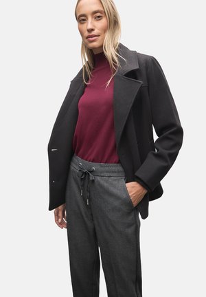 Zwarte jas met grote revers, bordeauxrode coltrui en grijze broek met trekkoord, met een subtiel patroon en zijzakken.