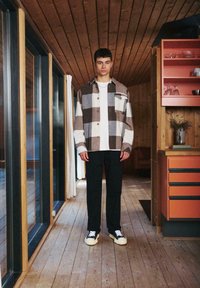 Men's flannel shirt with a brown, beige, and white check pattern, paired with black pants and white sneakers, in a wooden interior setting.