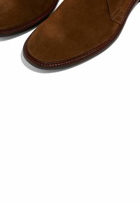Scarpe in suede marrone con texture liscia, punta arrotondata e cuciture borgogna a contrasto lungo la suola.