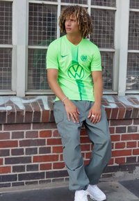 Maglia Nike verde neon con logo VW, abbinata a pantaloni cargo grigi. Il modello è appoggiato a un muro di mattoni, indossando sneakers bianche.