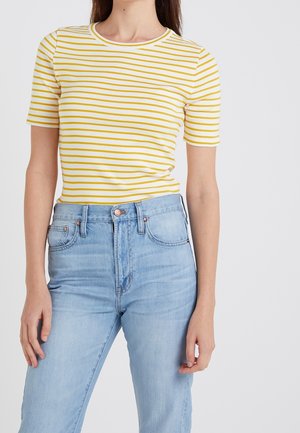 Kobieta ubrana w żółto-białą koszulkę w paski z krótkim rękawem, wpuszczoną do wysokich jasnoniebieskich dżinsów na jednolitym tle.