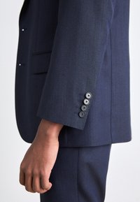 Giacca da abito blu navy in tessuto texture, con spacco singolo e polsini con quattro bottoni neri. Vestibilità su misura e finitura liscia.