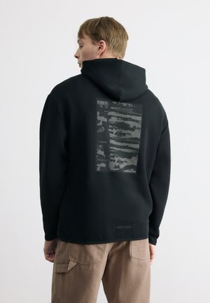 Młody mężczyzna zwrócony bokiem, ubrany w czarną bluzę z kapturem z prostokątnym abstrakcyjnym szarym nadrukiem na plecach oraz jasnobrązowe spodnie.