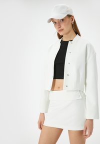 Witte cropped jacket met drukknoopjes, gecombineerd met een zwarte crop top en een witte rok. Het model draagt een witte cap en toont een gladde textuur.