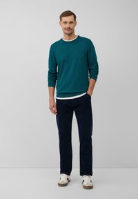 Mann trägt einen türkisfarbenen Pullover, ein weißes Unterhemd, dunkle blaue Hosen und weiße Turnschuhe und steht vor einem schlichten grauen Hintergrund.