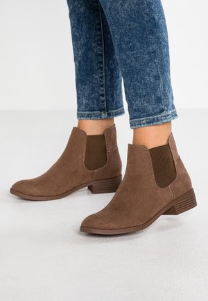Bottines en daim marron avec panneaux latéraux élastiques, portées avec un jean bleu retroussé sur un fond de sol clair.