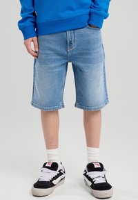 Vaaleansiniset farkkushortsit suoran leikkauksen, viimeistellyn helman ja sivutaskujen kanssa, yhdistettynä mustavalkoisiin lenkkareihin ja valkoisiin nilkkasukkien kanssa.