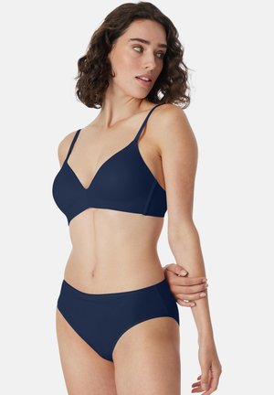 Donna con capelli ricci che indossa un reggiseno blu navy e mutandine abbinate, guarda di lato, tenendo un braccio con la mano opposta.