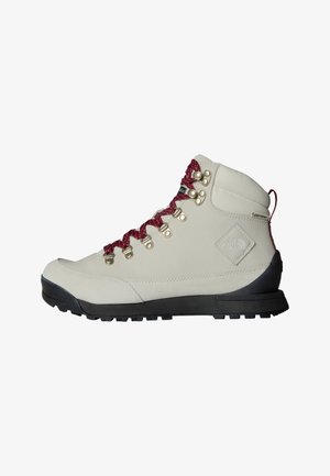 Bota de caminhada bege com cadarços vermelhos, ilhós de metal e uma superfície texturizada. Apresenta uma sola preta robusta e um detalhe de logotipo distinto na lateral.