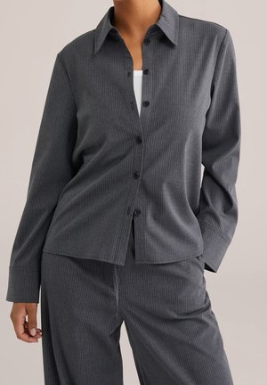 Vrouw die een donkergrijs verbrijzeld gestreept overhemd met knopen en bijpassende broek draagt met een wit topje eronder, staand tegen een effen achtergrond.
