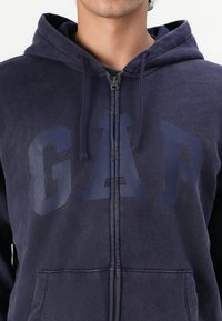 Sudadera con cremallera en azul marino hecha de un tejido suave, que presenta un gran logo de GAP en un tono más oscuro. Incluye capucha y bolsillos frontales.