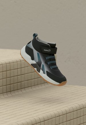 Zapatilla de caña alta negra y gris con detalles azules y cierre de velcro, colocada sobre escalones con baldosas beige contra una pared lisa.
