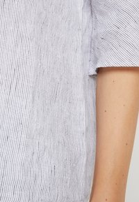 Bílá košile s krátkými rukávy a tenkými vertikálními černými pruhy nošená na paži s lehkou pletí a malými pihami.