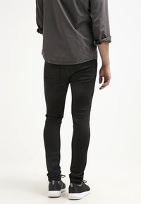 Tmavé úzké džíny s hladkou texturou a minimálním zdobením. Kombinovány s černými teniskami s bílými podrážkami. Viditelné zadní kapsy.