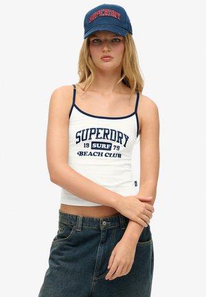 Witte tanktop met marineblauwe afwerking, voorzien van de opdruk "SUPERDRY BEACH CLUB"; gecombineerd met hoge denim shorts en een marineblauwe pet.