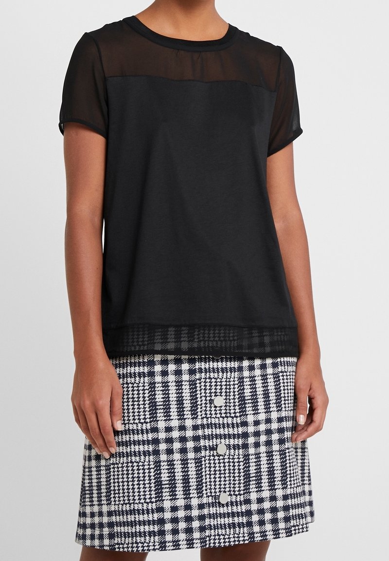 Camiseta negra con panel superior de malla transparente, mangas cortas; combinada con una falda de cuadros en blanco y azul marino con grandes botones y tejido texturizado.