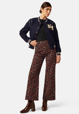 Vrouw draagt een navy varsity jack, zwart topje, bloementaillebroeken met wijde pijpen en bruine enkellaarzen, staande met hand op de heup.