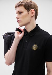 Melnā polo kreklā ar apkakli un zelta izšūtu logotipu. Audums šķiet gluds, ar šauru piegriezumu. Nes melnu somu.