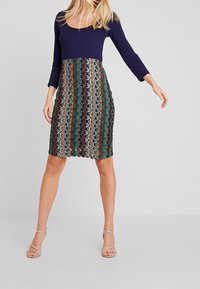 Sukienka z dopasowanym granatowym topem i wzorzystą spódnicą w geometryczne motywy w kolorach czerwonym, zielonym i kremowym; długość do kolan z teksturowanym wykończeniem.