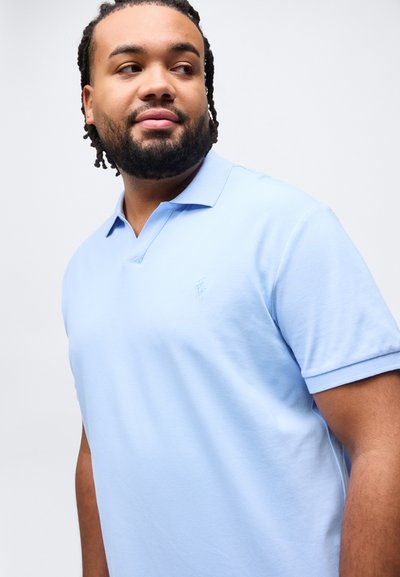 Άνδρας με πλεκτά μαλλιά και γενειάδα που φοράει ανοιχτό μπλε κοντομάνικο πόλο πουκάμισο, κοιτάζοντας προς τα αριστερά του μπροστά σε απλό φόντο.