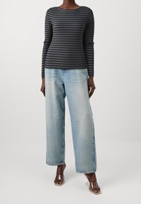 Langärmliges, dunkelgraues geripptes Oberteil mit horizontalen schwarzen Streifen, kombiniert mit hellblauen, weit geschnittenen Jeans und transparenten Sandalen.