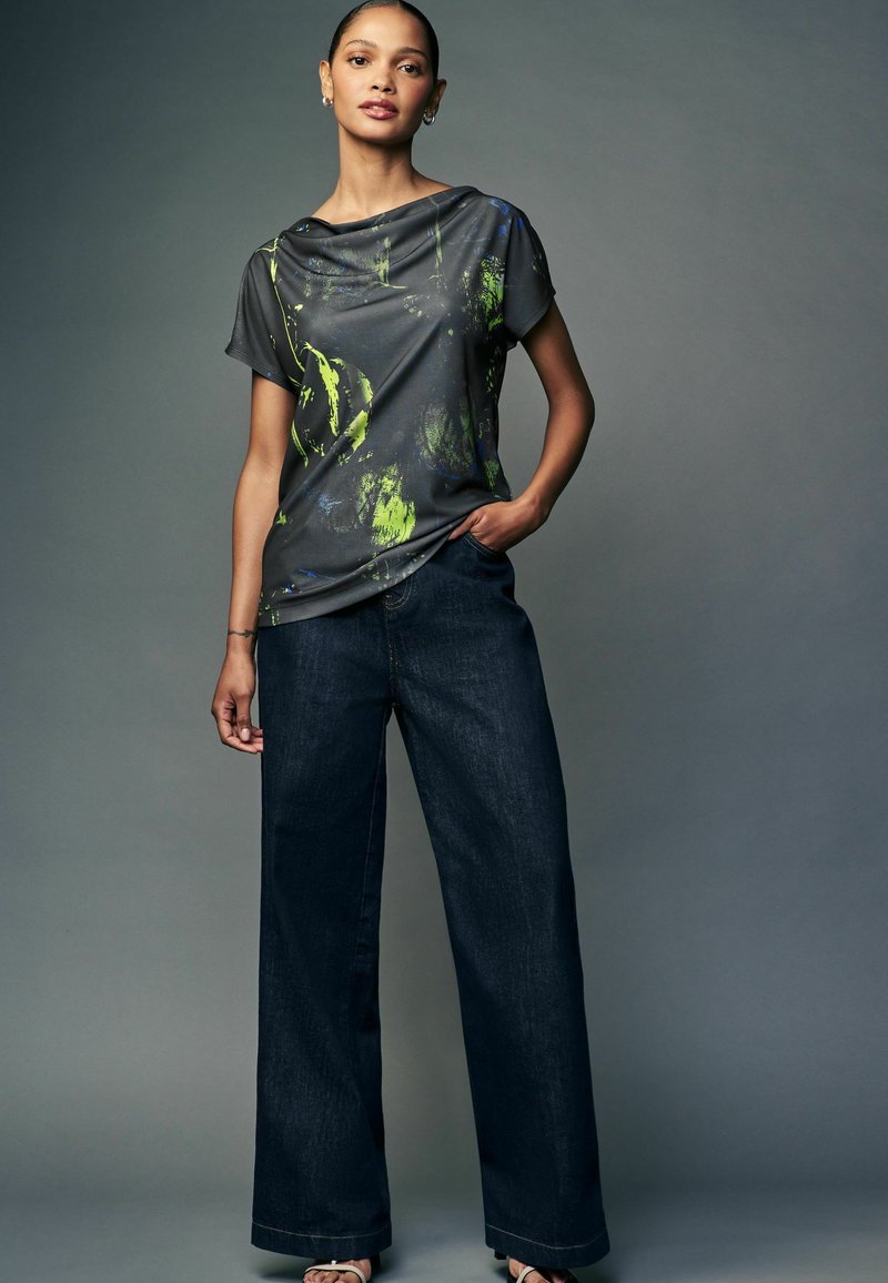 Cămașă gri cu un model abstract verde și albastru, combinată cu pantaloni din denim întunecați, cu croială largă. Design simplu, țesătură moale, aspect casual.