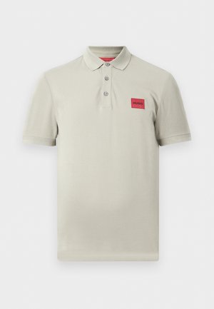 Polo beige clair à manches courtes avec trois boutons et un patch rectangulaire rouge marqué "HUGO" sur la poitrine gauche.