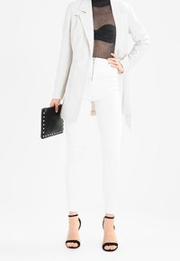 Biały, dopasowany żakiet na cienkim, czarnym przezroczystym topie, zestawiony z obcisłymi białymi, wysokim stanem jeansami i czarnymi sandałkami na obcasie z paskami, trzymając czarną kopertówkę z ćwiekami.