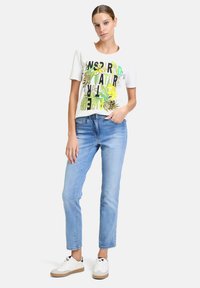 T-shirt blanche à manches courtes avec un texte graphique vert et noir et des motifs de feuilles, associée à un jean bleu clair ajusté et des baskets blanches.
