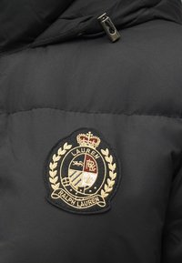 Fekete, párnázott dzseki, melyen egy Ralph Lauren hímzett címerfolt látható, amely koronát, babérkoszorút és osztott pajzsemblémát ábrázol.