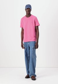 Ροζ βαμβακερό μπλουζάκι με στρογγυλή λαιμόκοψη, συνδυασμένο με ανοιχτό μπλε τζην παντελόνι cargo με πλαϊνές τσέπες και καφέ παπούτσια.