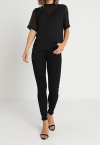 Top negro con mangas transparentes de puntos y frente sólido, combinado con jeans negros ajustados y tacones negros de tiras, mostrando un look monocromático.