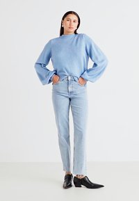 Lyseblå genser med lange, puffete ermer, kombinert med høyt liv, lyse blå jeans og spisse sorte ankelstøvler. Enkel bakgrunn.