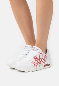 Vita Skechers-sneakers med ett röd droppande hjärta och "LOVE" grafitti som bärs på fötter mot en vit bakgrund.