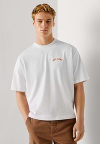 Weißes Baumwoll-T-Shirt mit kurzen Ärmeln, auf der linken Brust mit "PEPE JEANS" in geschwungener oranger Stickerei versehen. Einfacher und lockerer Schnitt.