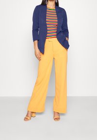 Blazer bleu marine, t-shirt rayé coloré et pantalon large orange. Tissu texturé, lignes épurées et sandales à talons carrés.