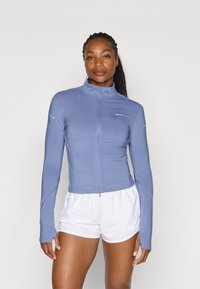 Long-sleeve zip-up jacket in light blue, featuring a high collar and smooth, stretchable fabric. Paired with white shorts.