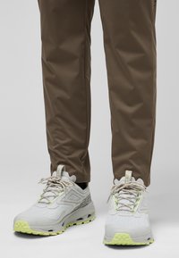 Lichtgrijze sportschoenen met een gestructureerde oppervlak, groene accenten en reflecterende veters. Draagt ze met een paar bruine broeken, wat een gestroomlijnd ontwerp laat zien.