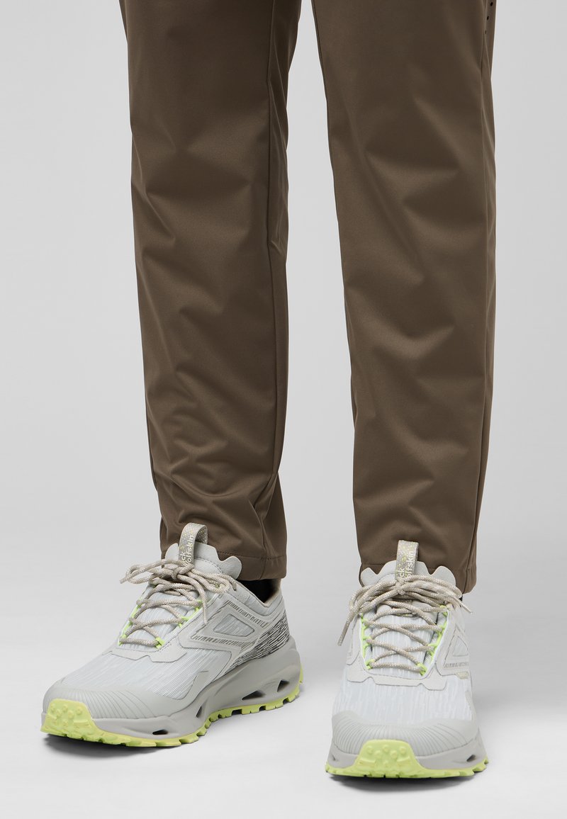 Lichtgrijze sportschoenen met een gestructureerde oppervlak, groene accenten en reflecterende veters. Draagt ze met een paar bruine broeken, wat een gestroomlijnd ontwerp laat zien.