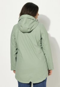 Grön vattentät jacka med huva, långa ärmar och rak skärning. Slät textur, har en baksidelinning och dragsko-detalj vid huvan.