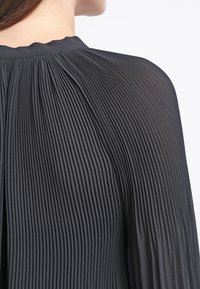 Czarna plisowana bluzka o teksturowanej powierzchni, z okrągłym dekoltem i marszczeniami z tyłu, które tworzą płynny ruch.
