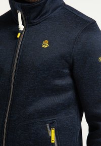 Námořnická modrá zapínací mikina vyrobená z texturovaného materiálu, která má vysoký límec, kapsy se žlutými akcenty a malým logem na hrudi.