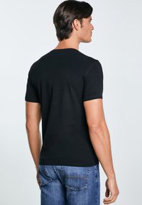 Mees seisab seljaga vaataja poole, kandes lihtsat musta lühikese varrukaga t-särki ja siniseid teksapükse, taustal on hele ühevärviline taust.