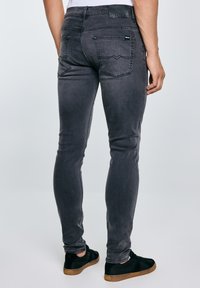 Rückansicht einer Person, die eng anliegende, ausgewaschene schwarze Jeans mit Gesäßtaschen und schwarze Sneaker mit braunen Sohlen trägt, stehend vor einem einfarbigen Hintergrund.