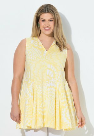 Smilende kvinde med blondt hår iført gul og hvid mønstret ærmeløs kjole, stående mod en ensfarvet hvid baggrund.