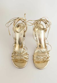 Sandales dorées à talons hauts avec plusieurs fines brides et longs lacets, vues de dessus sur un fond uni.