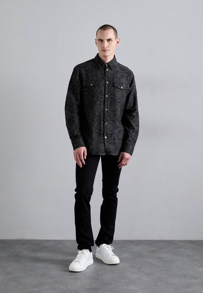 Chemise noire à manches longues avec un motif texturé, boutons sur le devant et deux poches poitrine, associée à un pantalon noir et des baskets blanches.