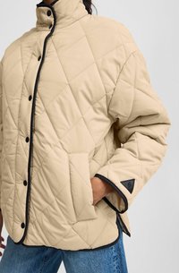 Veste beige matelassée avec des accents noirs. Présente un col haut, des boutons-pression et des poches latérales. Texture douce et détail de motif angulaire.